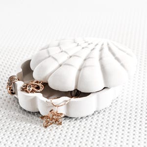 May include: A white, seashell-shaped jewelry box, partially open, revealing gold-colored rings and a star-shaped pendant necklace. The box has a textured surface and is set against a white, patterned background.