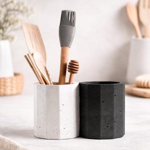 May include: Two geometric concrete kitchen utensil holders, one white and one black, with wooden cooking tools inside. The white holder contains a wooden spatula, spoon, and a gray silicone brush. A wooden honey dipper is also visible.
