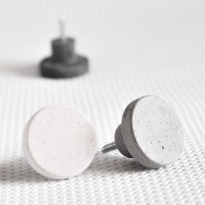 Puede incluir: Primer plano de dos pomos de armario de hormigón. Uno es blanco, el otro gris. Ambos tienen un diseño circular y un tornillo para la instalación. Un tercer pomo borroso es visible en el fondo. Estos pomos son ideales para muebles modernos. (Medidas en cm)