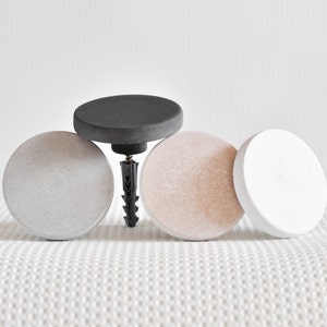 May include: Four round concrete wall hooks in grey, white and black. The hooks are attached to a white wall with a screw.