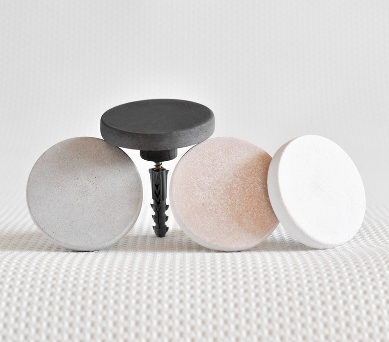 May include: Four round concrete wall hooks in grey, white and black. The hooks are attached to a white wall with a screw.