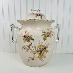 May include: A vintage, light pink ceramic jar with a lid, featuring floral designs in purple, yellow, and green. The jar has two handles and gold trim. The lid has a small handle. The jar is a decorative piece.
