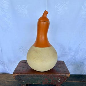 May include: A ceramic gourd-shaped vase with a lid. The vase is painted in two colors: orange and white. The vase is sitting on a wooden stool.