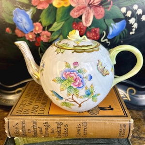 May include: A white porcelain teapot with a floral design and a butterfly. The teapot has a green lid and a gold trim. The teapot is sitting on top of two books, one of which is titled "Alice's Adventures in Wonderland and Through the Looking Glass by Lewis Carroll".