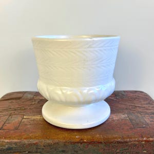 May include: A white ceramic planter with a pedestal base and a woven pattern around the top. The planter has a slightly off-white color.