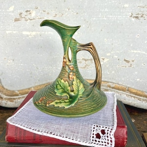 May include: Green ceramic pitcher with a leaf and berry design. The pitcher has a curved spout and a handle with a brown finish. The base is round and sits on a white lace doily over a red book.