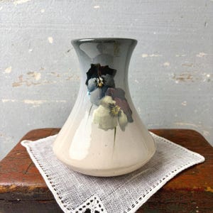 May include: A ceramic vase with a light gray and white finish, adorned with painted floral designs in shades of blue, purple, and green. The vase has a flared base and a slightly tapered neck. It sits on a white, lacy doily.