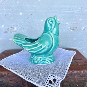 May include: A turquoise ceramic bird figurine with an open beak and a small opening on its back. The bird is perched on a white, lacy doily. The figurine has a glossy finish and is set against a weathered, light blue background.