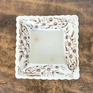 May include: A white square ceramic dish with a scalloped edge and a brown floral pattern around the rim.