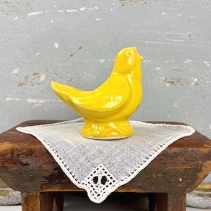 May include: A bright yellow ceramic bird figurine with a smooth, glossy finish. The bird is depicted in a perched position with its head turned slightly upward, featuring molded wing details and a rounded tail, resting on a small, textured base.