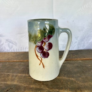 May include: A vintage ceramic mug with a hand-painted design of a bunch of grapes in shades of purple and green. The mug has a white background with a light blue glaze.