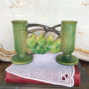 May include: Green ceramic vase with two cylindrical openings, textured sides, and a round base. A decorative element with green leaves and brown branches connects the vases. The vase is placed on a white lace doily.