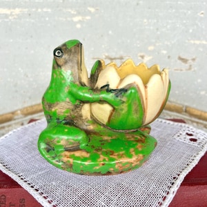 May include: A ceramic frog figurine in shades of green and brown, holding a cream-colored lotus flower. The frog is perched on a lily pad, with a distressed, vintage appearance. The flower's interior is yellow, adding a pop of color to the whimsical design.