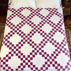 May include: A handmade quilt with a white background and a repeating pattern of burgundy and white squares. The quilt is spread across a bed with white pillows. The design creates a checkerboard effect, adding a touch of classic charm to the bedding.