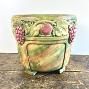 May include: A ceramic planter with a green, yellow, and brown painted finish. The planter features relief carvings of grapes, leaves, and an apple. The planter has a square base and a rounded top.