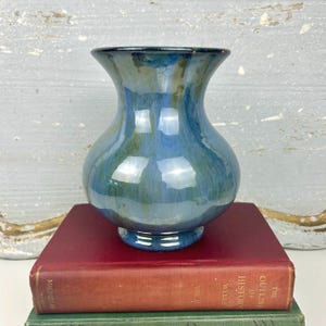 May include: A blue and green glazed ceramic vase with a flared rim and a bulbous body. The vase sits on a small pedestal base and reflects light. The vase is displayed on top of a stack of books.