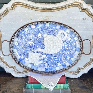 May include: Oval serving tray with a mosaic design featuring a blue and white pattern and a white teapot and cup design. The tray has a bronze-colored metal frame and handles. The tray is sitting on top of a stack of books.
