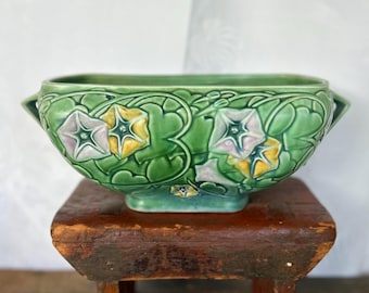 May include: A green ceramic planter with a square base and two handles. The planter is decorated with a floral design of pink and yellow morning glories.