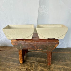 May include: Two white ceramic planter pots with a textured surface, sitting on a rustic wooden stool. The pots are rectangular with rounded edges and have a slightly raised base.