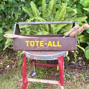 May include: A brown metal tool box with a handle and the yellow text TOTE-ALL printed on the front. The box contains two wooden-handled tools. The metal surface shows signs of wear and age, with a rectangular shape and an open top design.