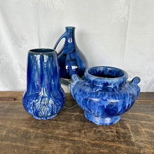 May include: Three blue ceramic vases with a mottled glaze. The vases are all different shapes and sizes. The largest vase is a round bowl with two handles. The medium vase is a tall, slender vase with a narrow neck. The smallest vase is a short, squat vase with a wide mouth.