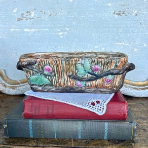 May include: A rectangular ceramic planter with a textured, bark-like exterior, adorned with painted branches, pink flowers, and green leaves. The planter sits atop a stack of vintage books, with a white lace doily between the planter and the books.