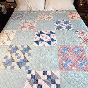 May include: A patchwork quilt featuring a grid of alternating solid light blue squares and patterned blocks. The patterned blocks display various geometric designs in shades of blue, red, white, and yellow, all finished with a diamond-shaped quilting stitch.