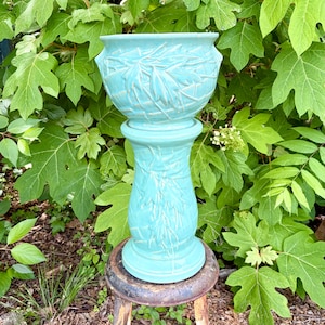 May include: A two-piece turquoise ceramic planter set consisting of a wide, bowl-shaped pot resting on a tall, cylindrical pedestal base. Both pieces feature a raised, textured leaf pattern with a glossy finish and integrated side handles.