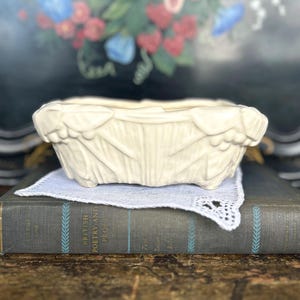 May include: A cream-colored rectangular planter with a textured, leaf-like design. The planter sits on a white lace doily atop a vintage book. The background features a blurred floral arrangement, suggesting a vintage or antique aesthetic.