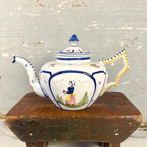 May include: White ceramic teapot with blue and yellow accents. The teapot features a hand-painted design of a woman in traditional clothing, flowers, and foliage. The handle is yellow with blue dots.