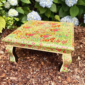 May include: A square, hand-painted wooden table with a floral design in shades of green, pink, red, and yellow. The table has curved legs and is set outdoors. The table is approximately 12 inches long, 12 inches wide, and 6 inches tall.