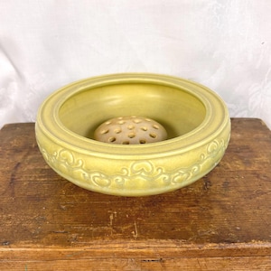May include: A yellow ceramic bowl with a decorative design and a small, round, beige flower frog in the center.