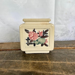 May include: A small, square, cream-colored ceramic planter with a floral design. The planter features pink roses, green leaves, and brown branches.
