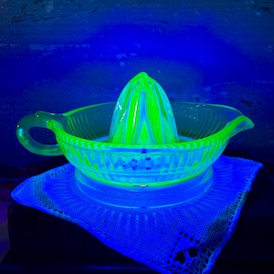 May include: A vintage, neon green glass citrus juicer with a conical reamer and a bowl with a handle and spout. The juicer sits on a white lace doily, illuminated by a blue light, creating a striking contrast.