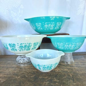May include: Set of four vintage turquoise and white mixing bowls with a folk art design. The bowls are nested inside each other, with the largest bowl on the bottom and the smallest bowl on top.