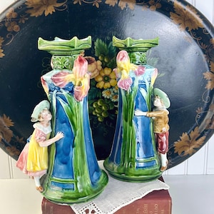 May include: Pair of vintage ceramic vases with a floral design and figures of children. The vases are blue, green, and pink, with a detailed iris flower design. The children are wearing hats and are hugging the vases.