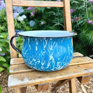 May include: A blue and white enamelware bowl with a black handle and rim. The bowl has a marbled pattern and sits on a wooden chair. The background features green foliage and purple flowers.