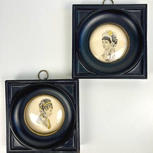 May include: Two framed portraits featuring vintage illustrations of women in profile. The portraits are set in black square frames with circular openings, and each has a small metal loop for hanging. The illustrations are in muted tones.