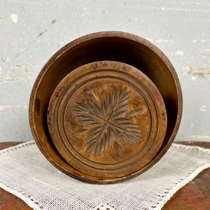 May include: A round, brown wooden butter mold featuring a carved leaf pattern on the circular lid. The mold has a deep, cylindrical body with a natural wood grain finish and visible signs of wear, showing a dark, aged patina throughout the surface.