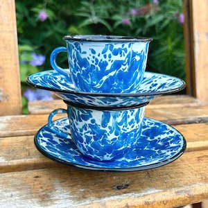 May include: Two stacked blue and white enamelware coffee cups and saucers. The cups and saucers have a speckled pattern with a black rim. The cups are sitting on a wooden surface with a blurred green background.