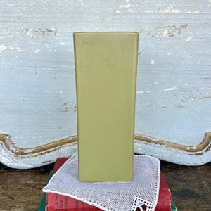May include: A tall, rectangular, olive-green vase. The vase is placed on a stack of books and a white doily with a decorative edge. The background features a distressed, light blue wooden surface.