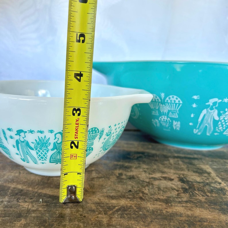 May include: Two white mixing bowls with a turquoise blue rim and a white and turquoise blue pattern of people and flowers. The smaller bowl is 7 inches in diameter.
