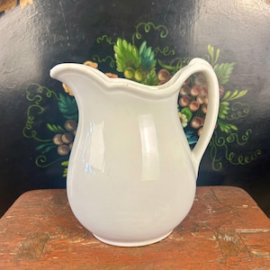 May include: A white ceramic pitcher with a curved handle and a scalloped rim. The pitcher is set against a dark background with a painted floral design and rests on a weathered wooden surface.