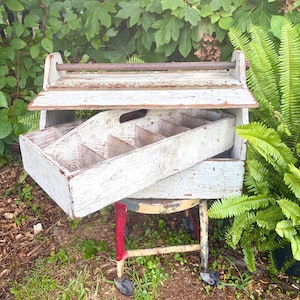 May include: A weathered white wooden tool caddy with a hinged lid and a dark metal handle. The caddy features an interior divided into multiple rectangular compartments for organization, showing visible wood grain and distressed paint finish.