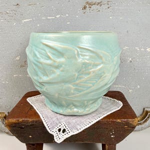 May include: A light blue ceramic planter with a textured, embossed design of leaves and vines. The planter sits on a small, dark wooden table with a white, lacy doily. The planter has a rounded shape and a slightly flared base.