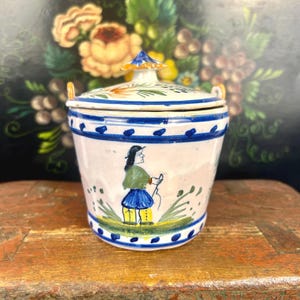 May include: A hand-painted ceramic jar with a lid. The jar is white with blue and yellow accents, featuring a painted figure in a green top and blue skirt. The lid has a floral design and a blue finial. The jar has two small handles.