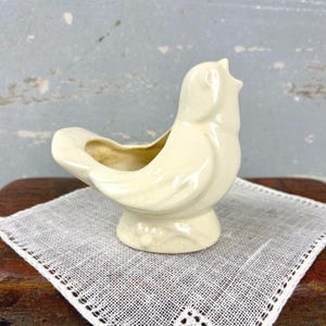 May include: A cream-colored ceramic bird-shaped planter with a glossy finish and a subtle crackle glaze texture. The bird is depicted with its head tilted upward, featuring a hollowed-out back section for holding small items or plants.