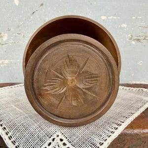 May include: A round, dark brown wooden container with a removable lid. The lid features a carved, four-petaled floral design in the center. The container has a smooth, polished finish and a deep, hollow interior, showcasing the natural wood grain.