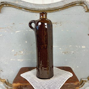 May include: A tall, cylindrical ceramic bottle with a dark brown, glossy glaze. The bottle features a single curved handle attached near the narrow neck and a smooth, reflective surface that catches the light, showcasing its deep, uniform color.