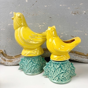 May include: Two ceramic bird-shaped planters, each featuring a bright yellow bird perched atop a textured teal green base that resembles a leafy tree stump. The birds have open beaks and molded wing details, while the bases have intricate foliage.